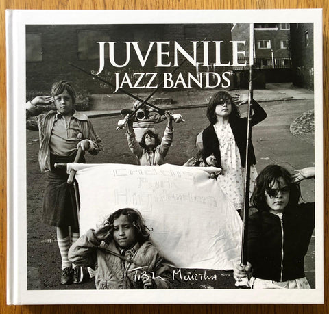 The photography book cover of Juvenile Jazz Bands by Tish Murtha. Hardback book in B&W with photography of children holding a sign.