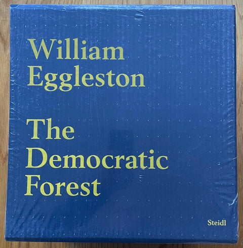 The photography book cover of The Democratic Forest by William Eggleston. Blue slipcased set of 10 hardbacks. Yellow writing.