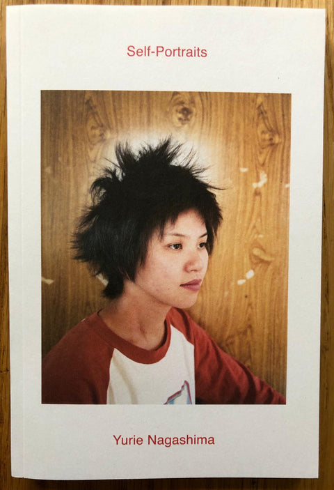 The photography book cover of Self-Portraits by Yurie Nagashima. Paperback in white with image of a person with spikey black hair standing in front of wooden wall.